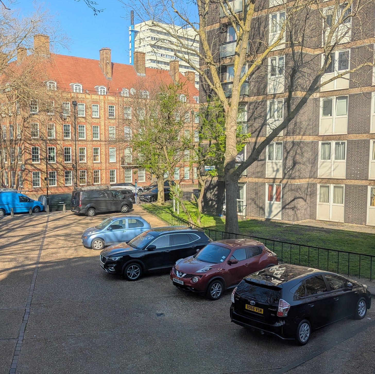 A car park on a sunny days, with a few cars parked, flanked by a tower block and a medium sized apartment building. A car park on a sunny days, with a few cars parked, flanked by a tower block and a medium sized apartment building.