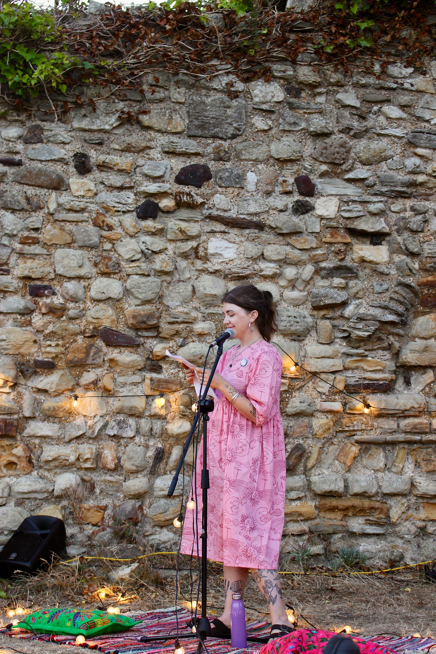 Grace is reading from A4 plain paper pages against a rocky castle ruins wall, in a pink smock dress covered in doodles. she's lit by fairy lights from underneath, and definitely isn't pregnant.