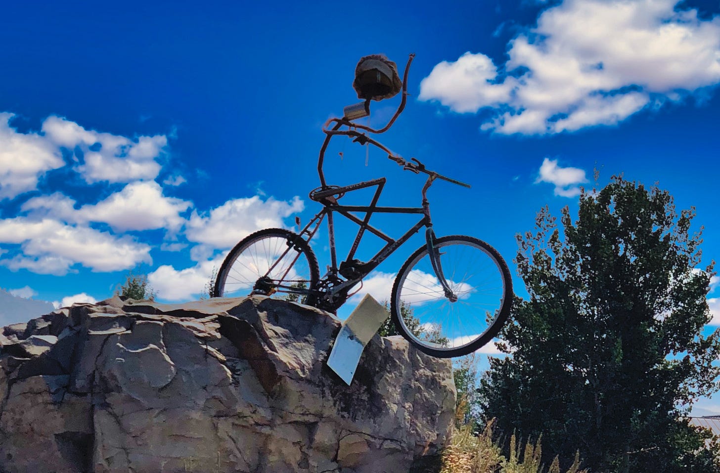 A sculpture made from an old bicycle with a metal rider with a cap and a notebook that has fallen on the rocks