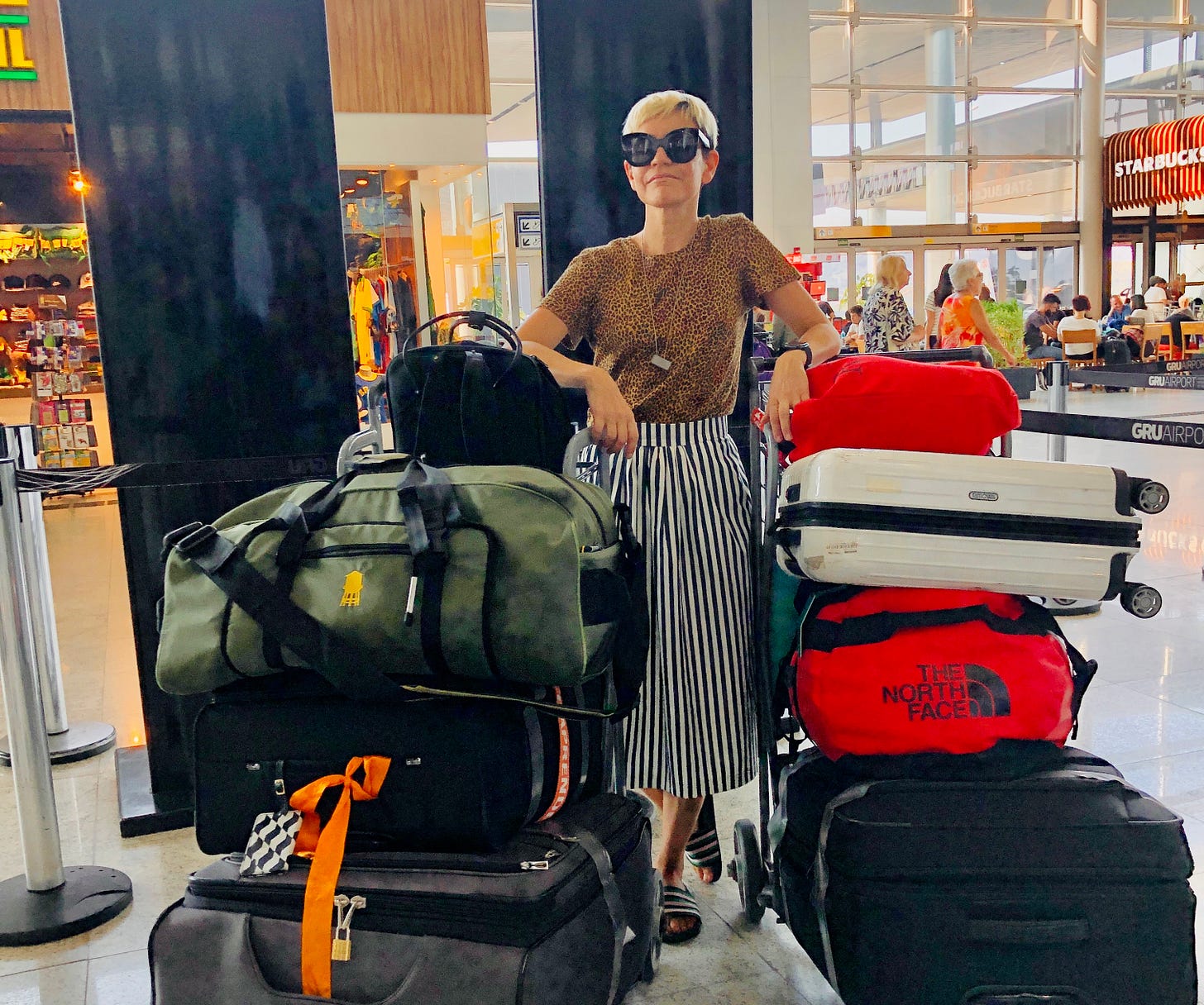 Pessoa em pé no aeroporto com muitos volumes de bagagem empilhados em dois carrinhos, usando óculos escuros e expressão confiante. Ao fundo, área de embarque movimentada com lojas e cafés. Pessoa em pé no aeroporto com muitos volumes de bagagem empilhados em dois carrinhos, usando óculos escuros e expressão confiante. Ao fundo, área de embarque movimentada com lojas e cafés.