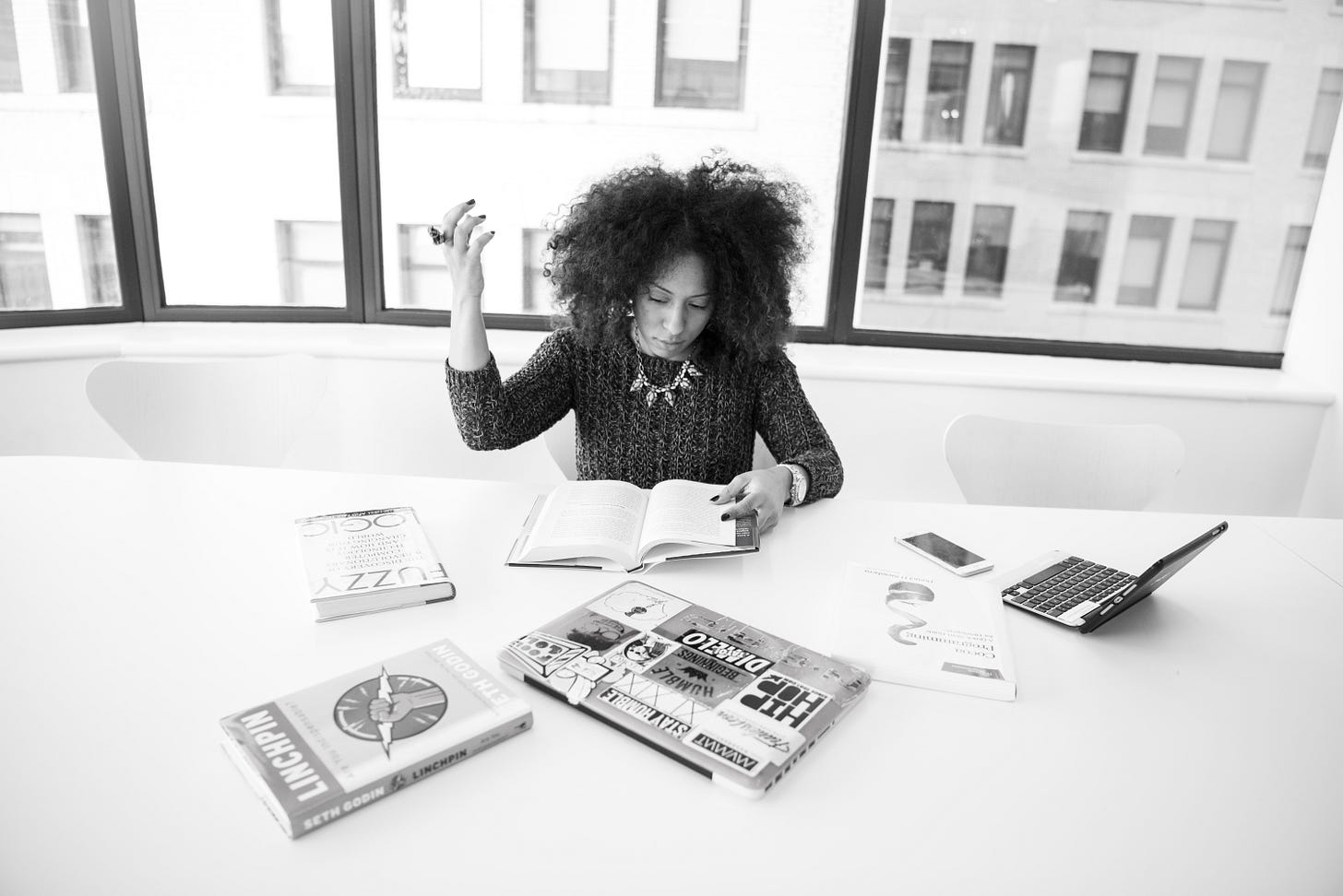 A woman sits at a desk covered with books and two laptops, plus a phone. She holds open one of the books and has her other hand up in the air, as if in frustration.