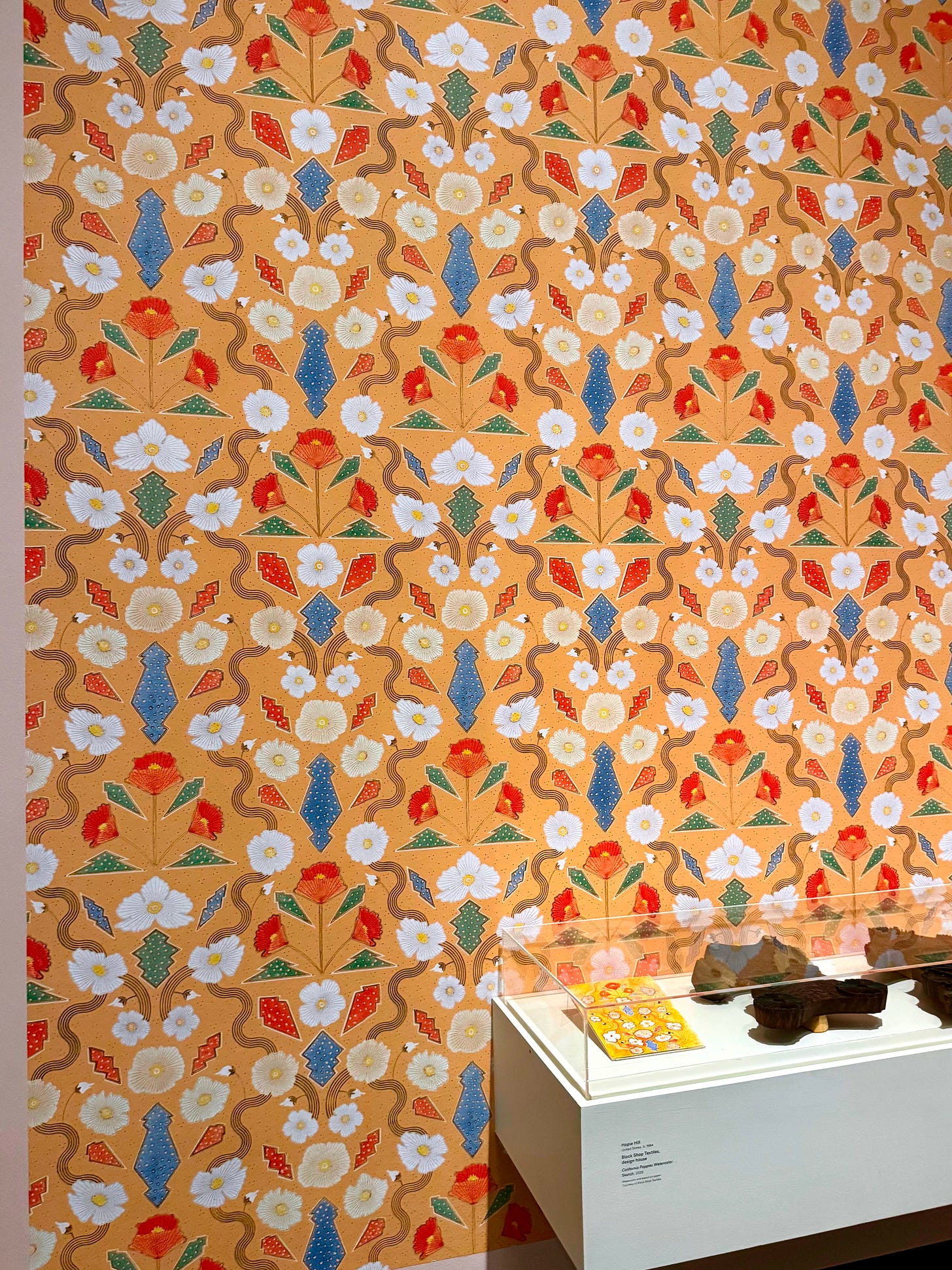 A gallery wall covered in a repeating floral wallpaper pattern in ochre, blue, orange, green, and white, with a display case positioned in front.