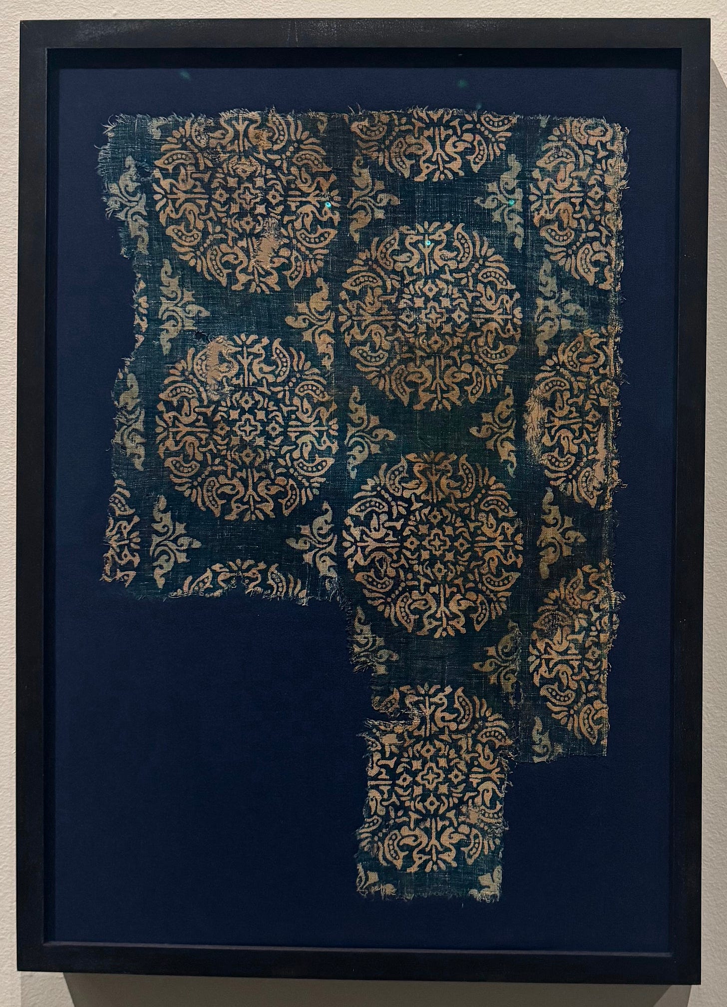 A historic indigo textile fragment patterned with repeating gold floral and botanical blocks, displayed in a dark frame.