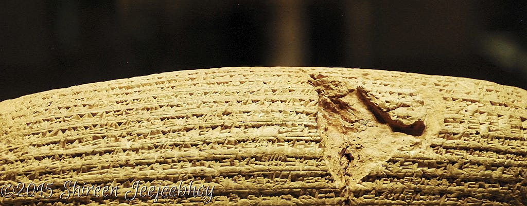 The Cyrus Cylinder. Shireen Anne Jeejeebhoy photo. Shot at the British Museum, 15 March 2015. Against a black background, the curvature and top half of the cylinder is highlighted by artificial lights, showing the cuniform in sharp relief. The Cyrus Cylinder. Shireen Anne Jeejeebhoy photo. Shot at the British Museum, 15 March 2015. Against a black background, the curvature and top half of the cylinder is highlighted by artificial lights, showing the cuniform in sharp relief.