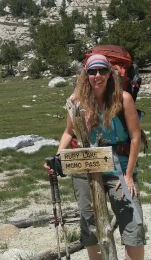 A photograph of Christie, who is seen standing with hiking poles in front of a river. She wears sunglasses, a bandana, and a hiking backpack. Before her is a sign that reads, "Ruby Lake / Mono Pass," with an arrow pointing forward.