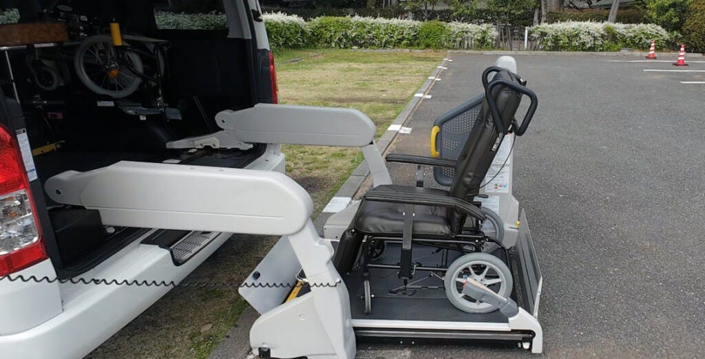A rear wheelchair lift extended from the back of an accessible taxi van, with a folding seat lowered onto the platform and a manual wheelchair stored inside the vehicle. A rear wheelchair lift extended from the back of an accessible taxi van, with a folding seat lowered onto the platform and a manual wheelchair stored inside the vehicle.