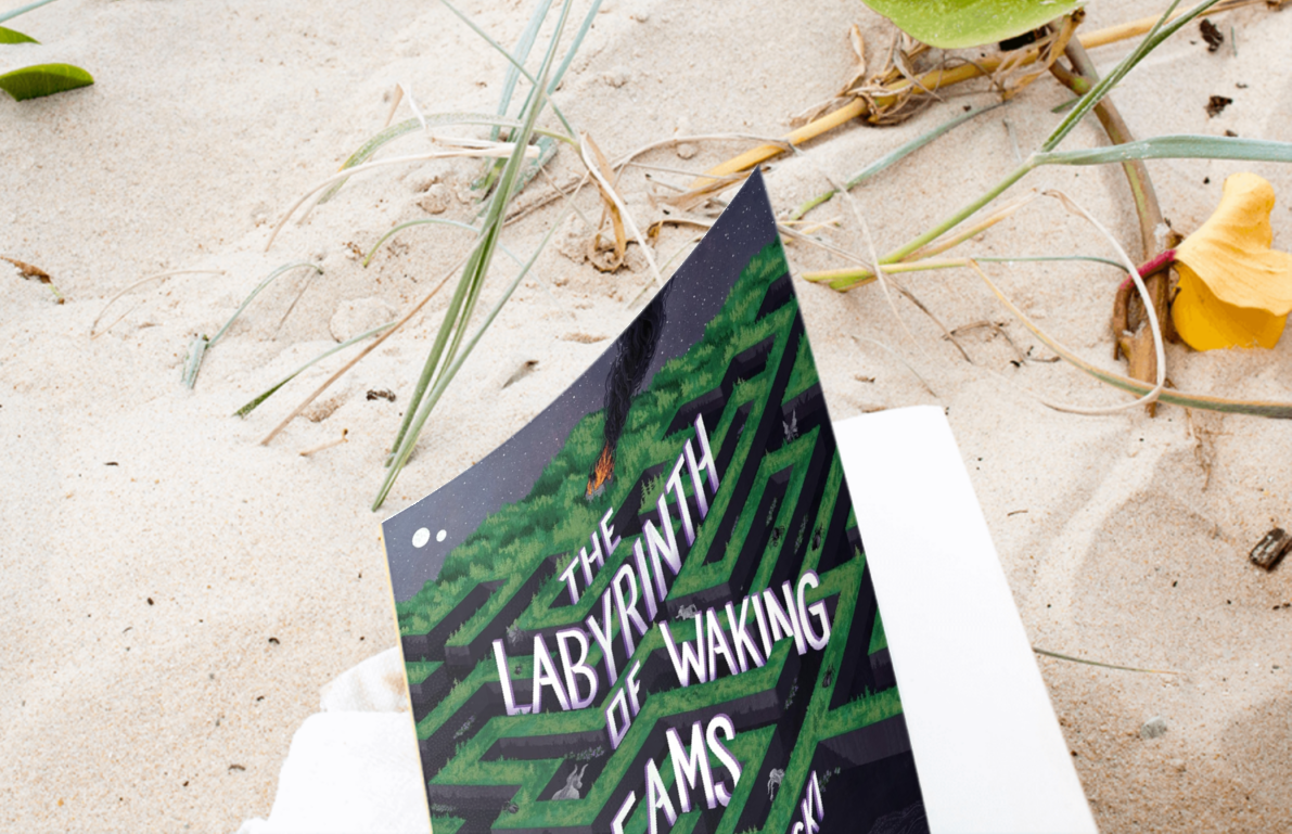Book "The Labyrinth of the Waking Dead" in the sand by a beach with foilage.