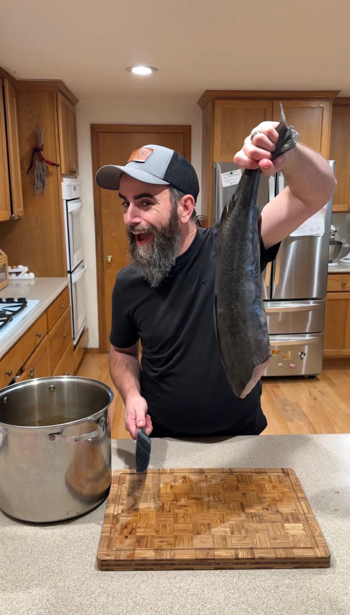 A man in a kitchen holds up a whole fish beside a cutting board and pot.