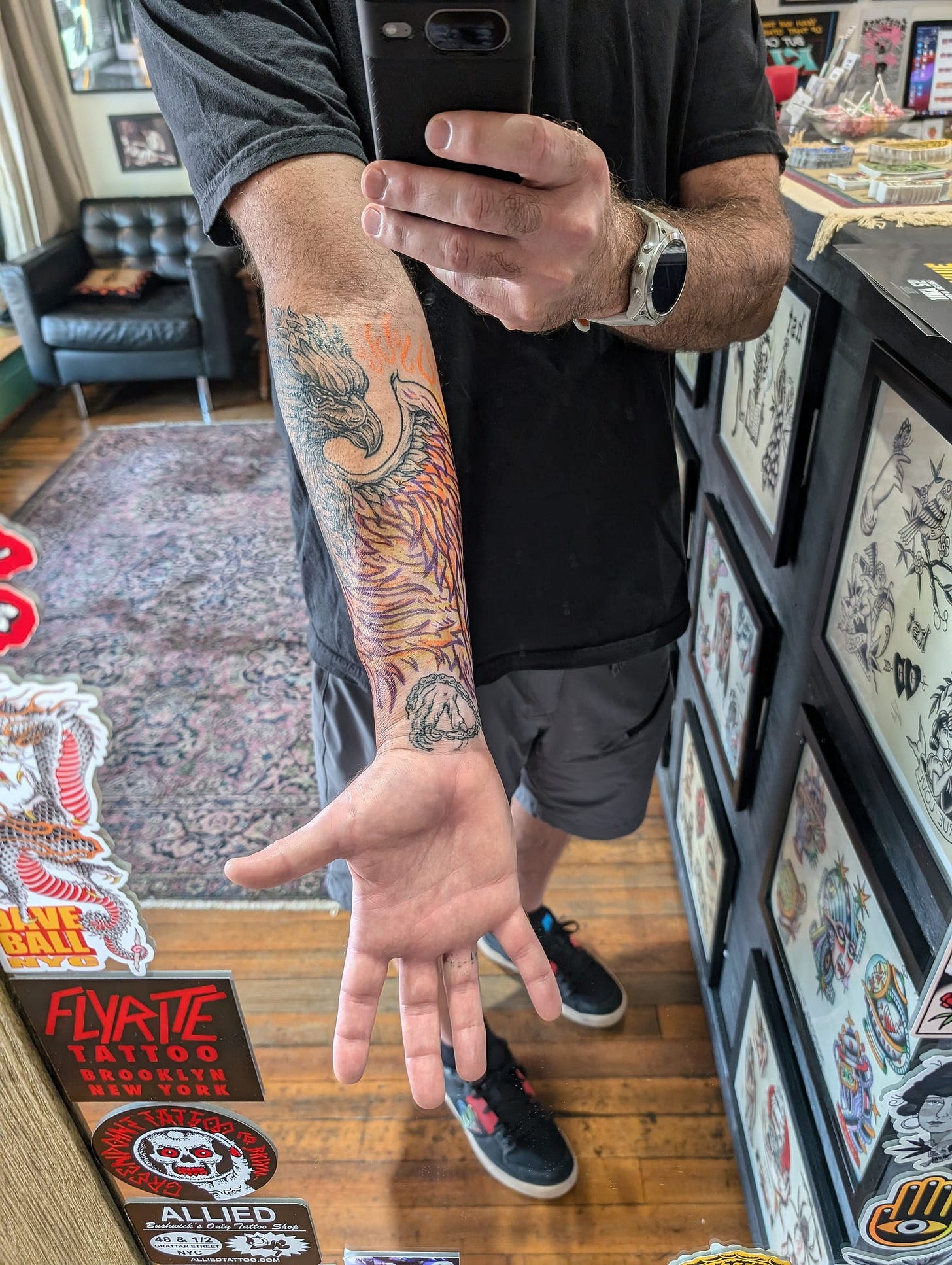 A photograph of the author in a mirror at a tattoo shop. He can see his reflection from his chest to his shoes while holding out his outstretched left arm. On his arm are completed tattoos, the head of a phoenix near the crease of his elbow and the claws on his wrist. A hand drawn stencil of the bird’s wing connects the tattoos. A photograph of the author in a mirror at a tattoo shop. He can see his reflection from his chest to his shoes while holding out his outstretched left arm. On his arm are completed tattoos, the head of a phoenix near the crease of his elbow and the claws on his wrist. A hand drawn stencil of the bird’s wing connects the tattoos.