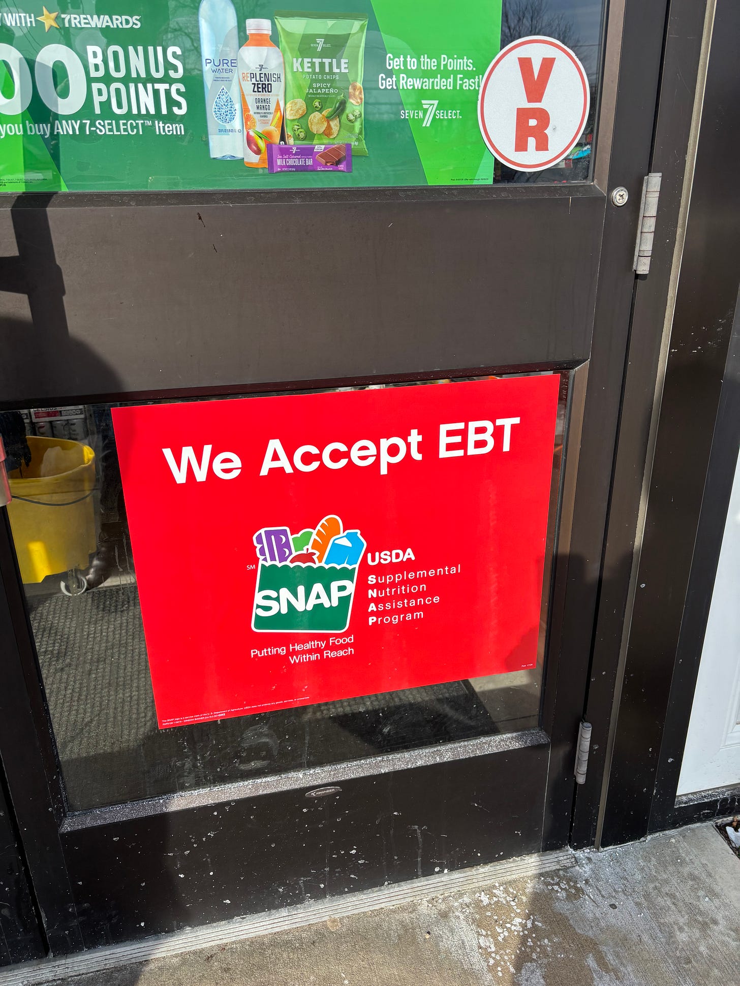 A close-up of a worn franchise convenience store front door featuring a prominent red "We Accept EBT SNAP" sign below a corporate advertisement for 7-Eleven rewards points and processed snacks.