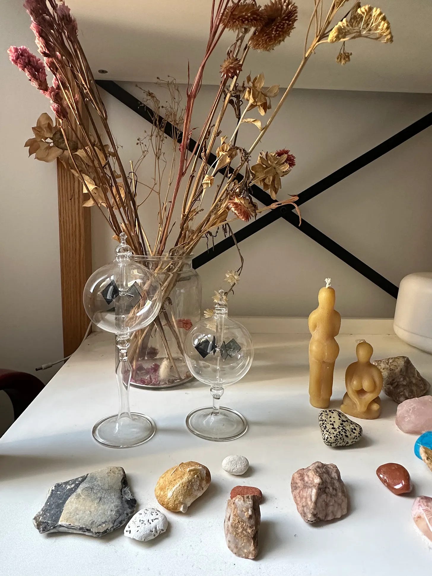 A tabletop arrangement with two glass light mills, dried flowers, sculptural candles, stones, shells, and small collected objects arranged near a vase. A tabletop arrangement with two glass light mills, dried flowers, sculptural candles, stones, shells, and small collected objects arranged near a vase.