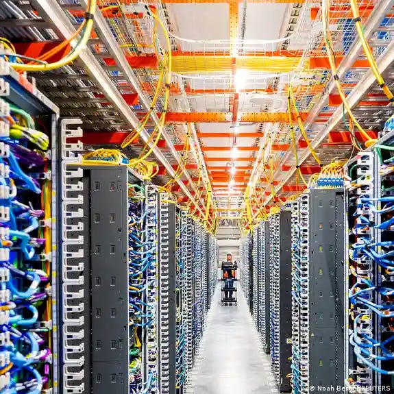 A person at the end of a long corridor of tall banks of computers, with a lot of blue wiring visible.