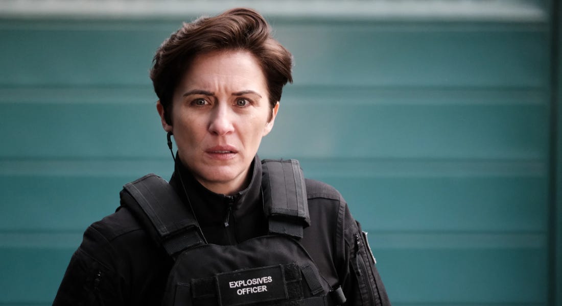 A woman - Vicky McClure - wearing a black jacket and vest that says 'explosives officer', with a blue background behind her A woman - Vicky McClure - wearing a black jacket and vest that says 'explosives officer', with a blue background behind her