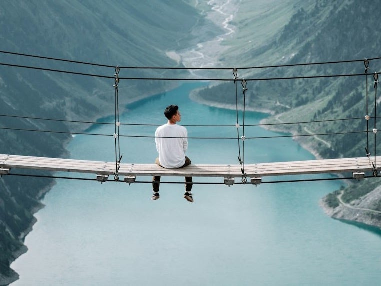 man wearing white shirt siting on bridge overlooking at mountain