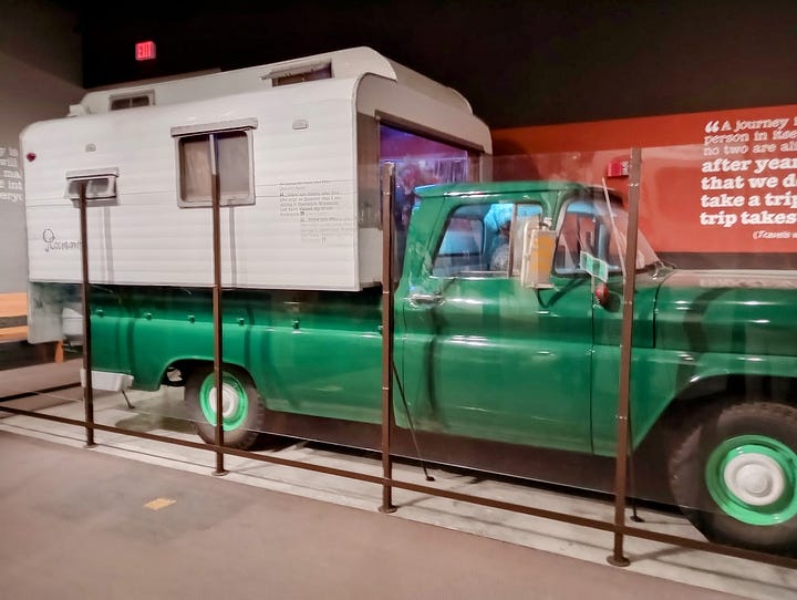 Pickup truck with camper shell, with Rocinante written on side, dog looking out of passenger window, narrow interior with typewriter on table and stove, and man with dog