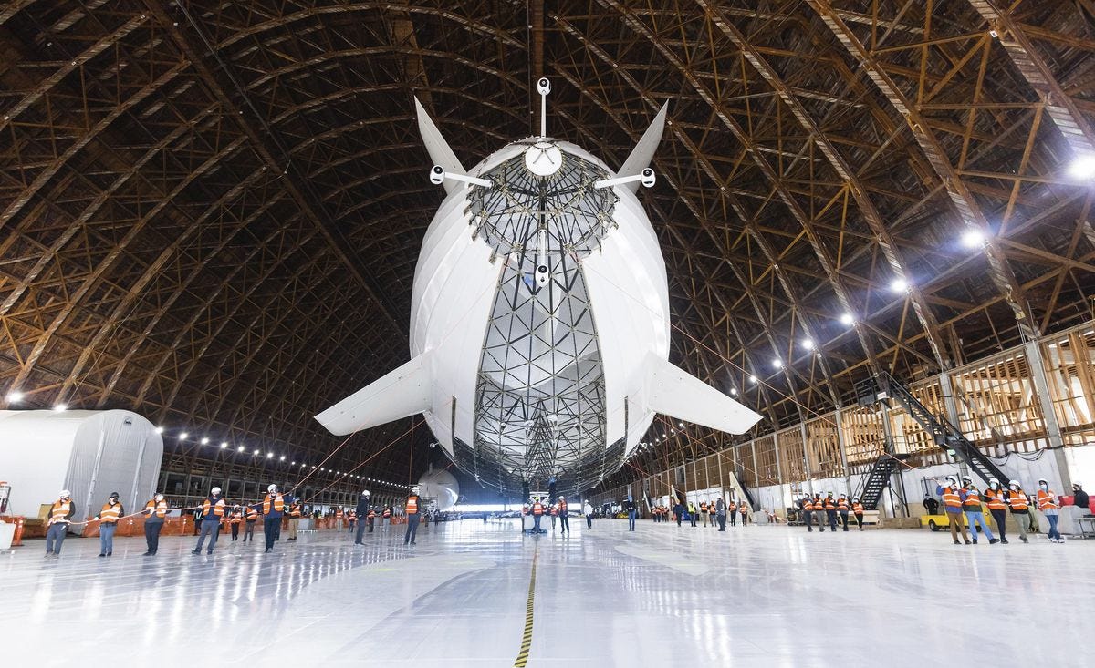 People are arranged around the rear end of a white, cylindrical airship above a white floor and inside of a large aircraft hanger.