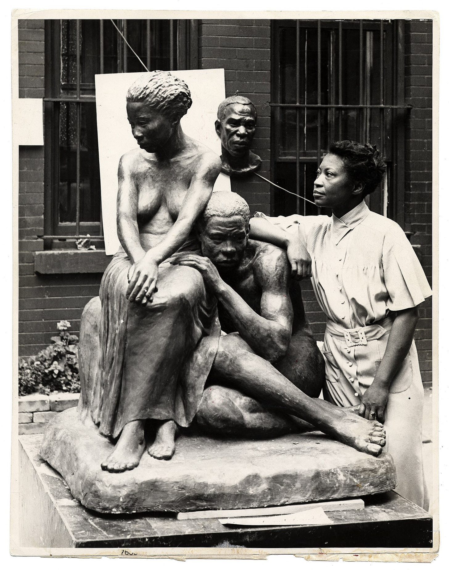Augusta Savage standing beside her plaster sculpture of a seated woman and a seated man, with a bust on a board behind them. Augusta Savage standing beside her plaster sculpture of a seated woman and a seated man, with a bust on a board behind them.