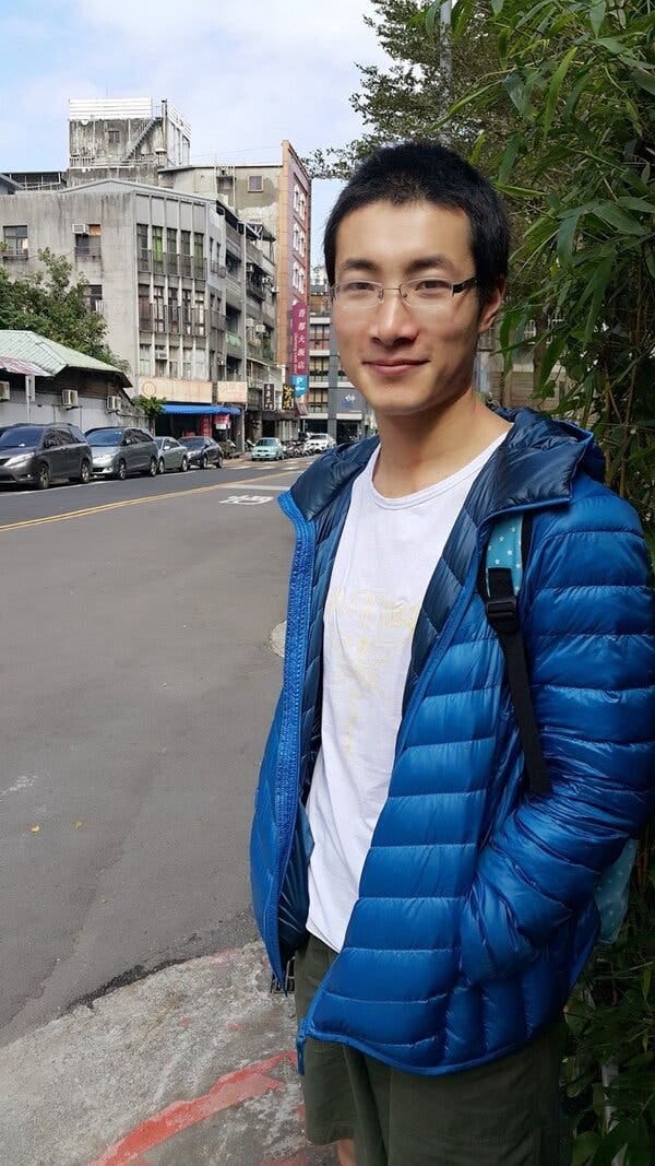 A young man, wearing a blue parka and a backpack, stands on a street, smiling.