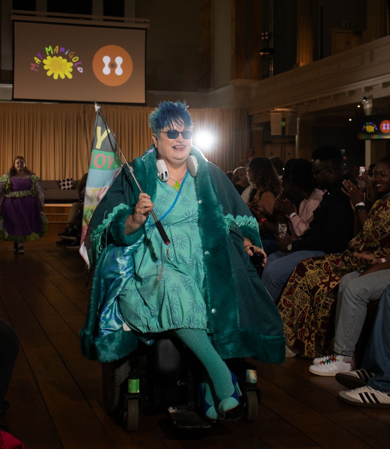model in a teal colored dress using a power wheelchair on the runway and waving a disability pride flag