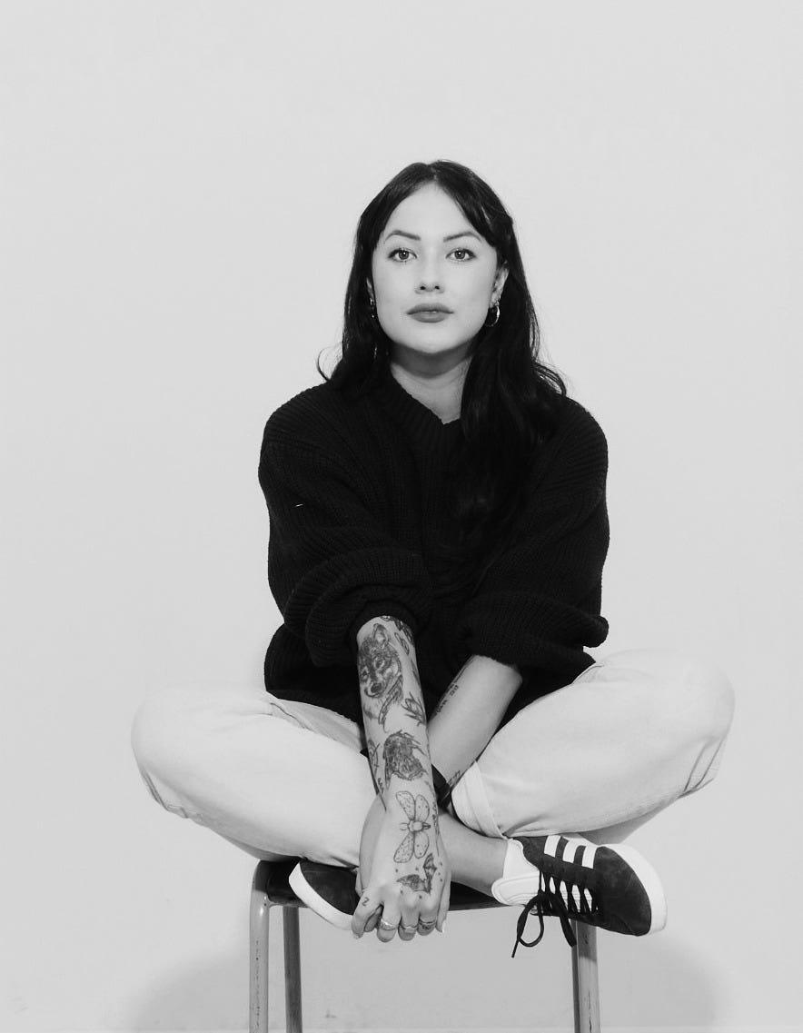 Black-and-white author portrait for a Tarot DMs interview, showing a woman seated cross-legged on a chair with tattooed arms, dark hair and a calm direct gaze against a minimal studio backdrop.