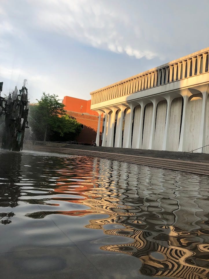 A view across rippling water, with an abstract fountain made of metal on the left, and a large white building on the right. A view across rippling water, with an abstract fountain made of metal on the left, and a large white building on the right.