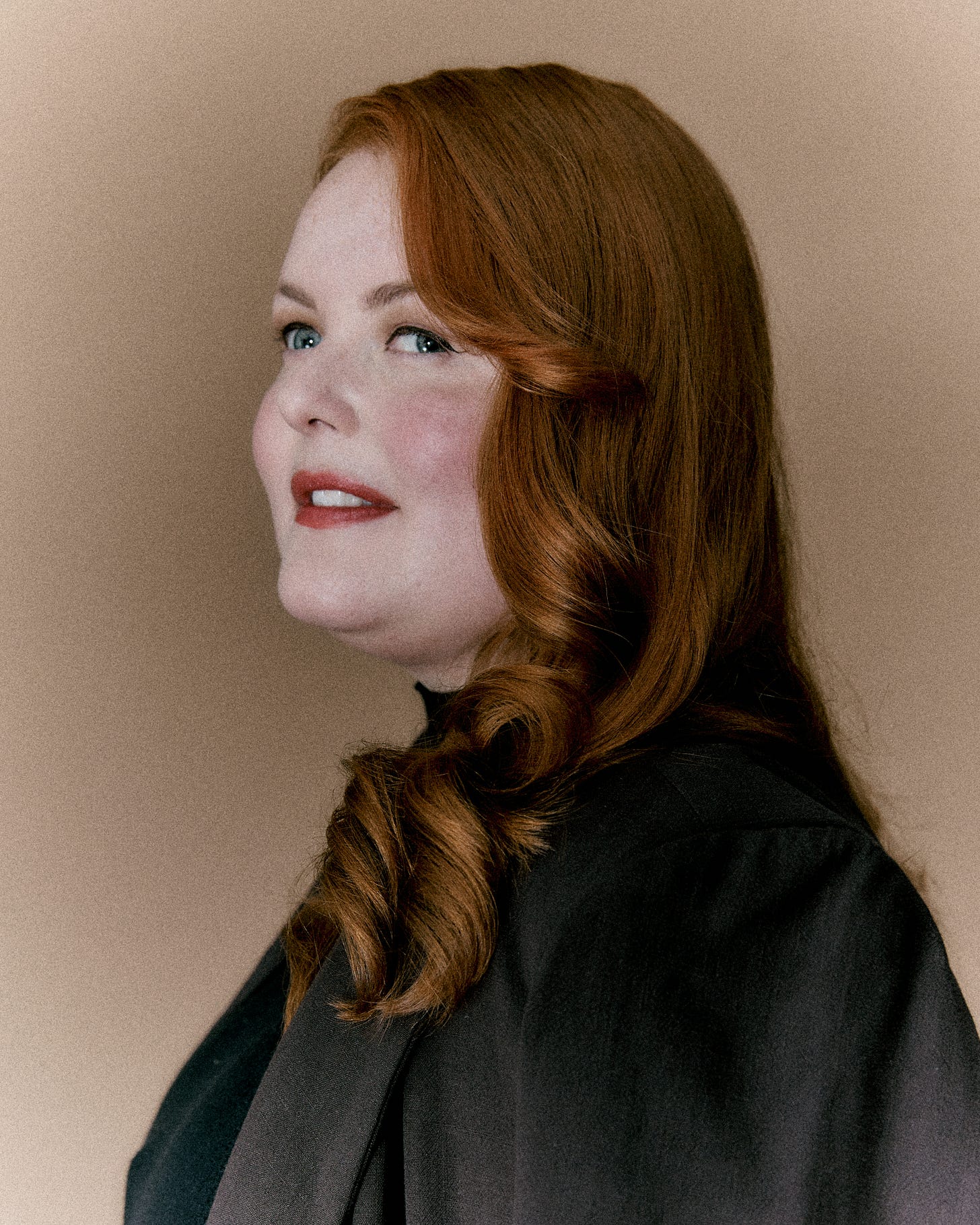 A full colour portrait of Lucy. The photo captures her side profile, highlighting her luminous pale skin and blue eyes lined with black eyeliner. Her red hair frames her face in full, glossy waves and she wears a slate grey blazer.