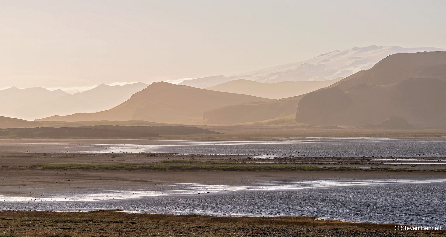 "Layered mountain silhouettes fade into haze above a tidal flat near Dyrhólaey, Iceland, in warm, muted light." "Layered mountain silhouettes fade into haze above a tidal flat near Dyrhólaey, Iceland, in warm, muted light."