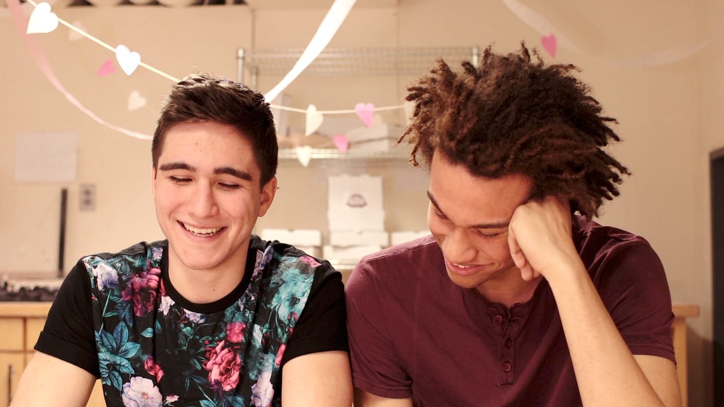 In this still from the Middlemarch web series, Fred Vincy (Oliver Shoulson) and Max Garth (Kai Nugent) sit side by side with their arms lightly touching. Both are smiling, and there are some heart-shaped decorations and stacked pizza boxes in the background.