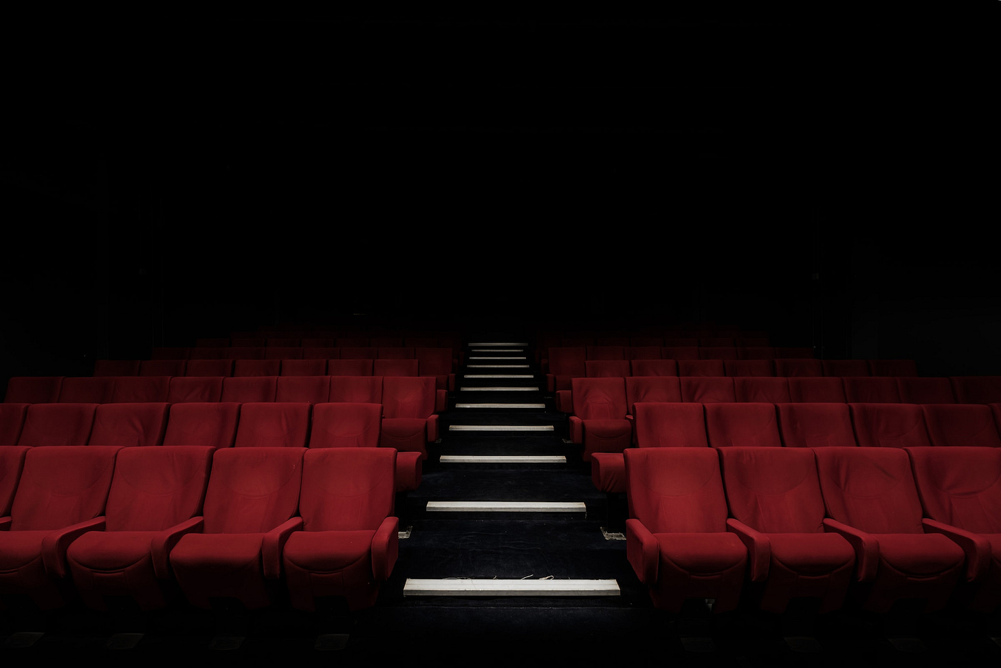 File di poltrone rosse del cinema viste dal davanti, con lo sfondo che sfuma verso il nero. Nella parte centrale scalini neri con bordo bianco che separano le due sezioni di poltrone.