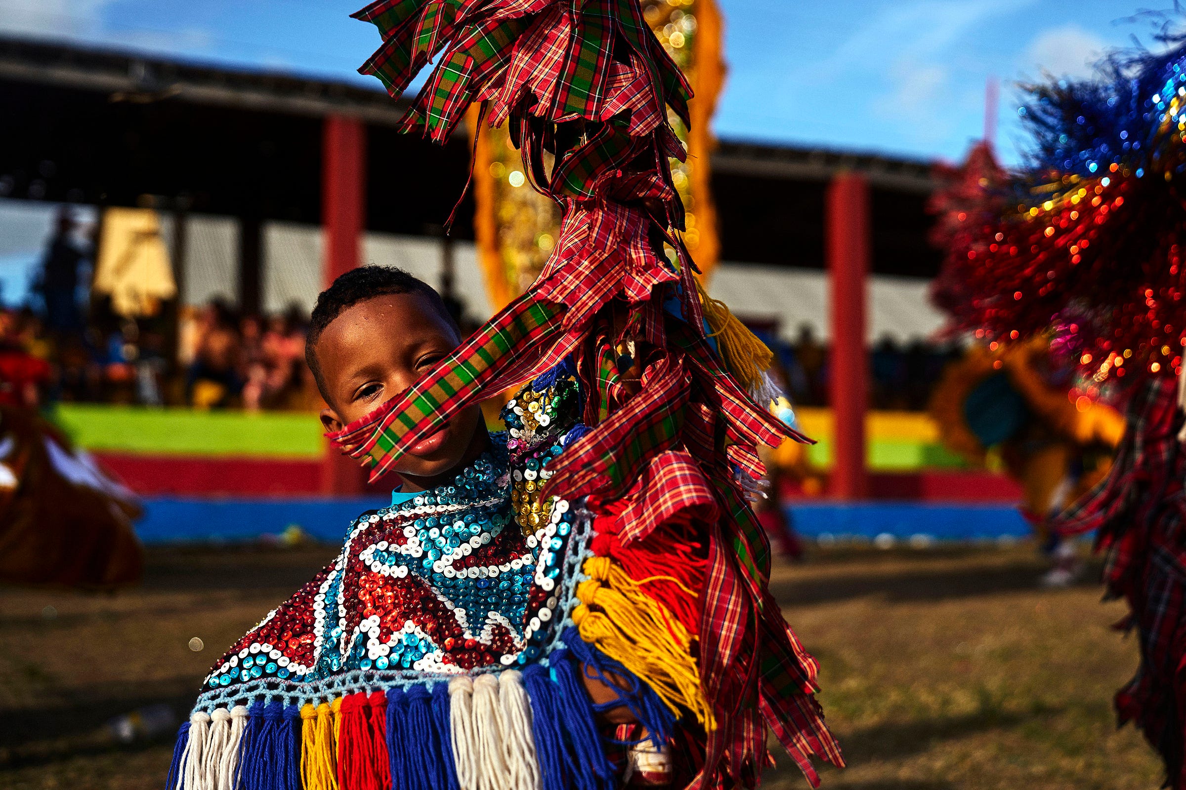 A child in a colorful, sequined costume with fringes and plaid ribbons dances outdoors during a vibrant cultural festival, with blurred spectators and bright decorations in the background.