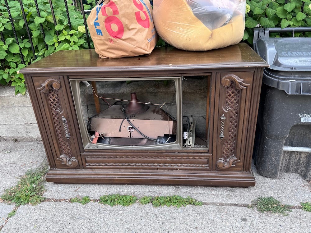 Smashed in TV console on a curb with trash bags and can.