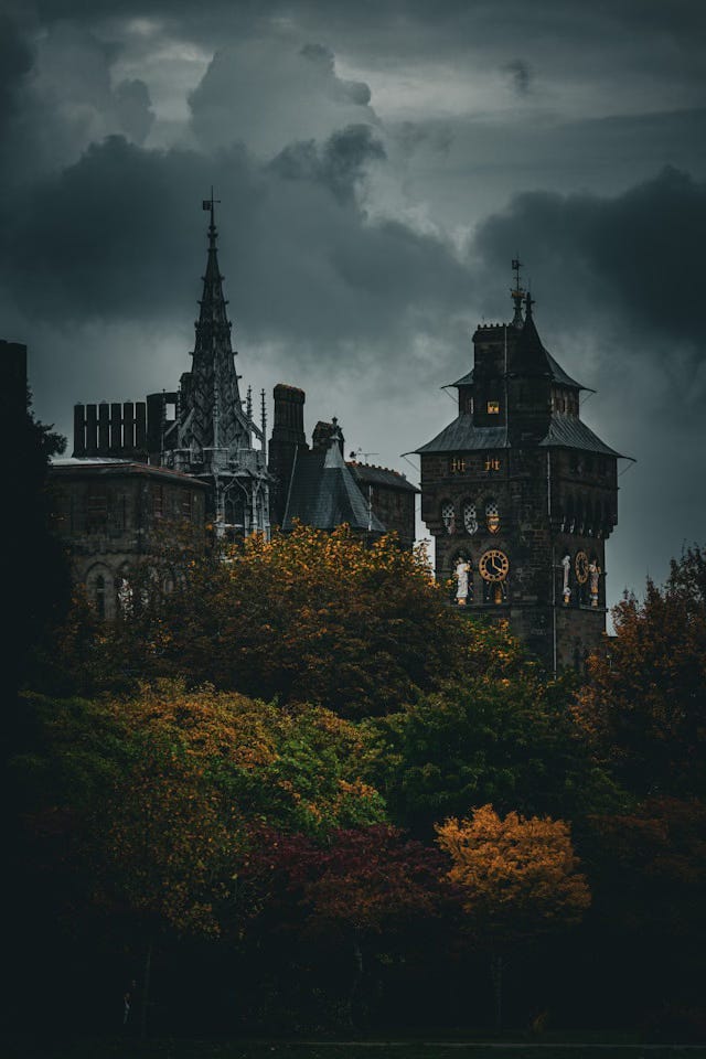 An elaborate grey stone castle with a turett and a clock tower, surrounded by dark green and gold foliage. Ominous dark clouds fill the sky overhead.