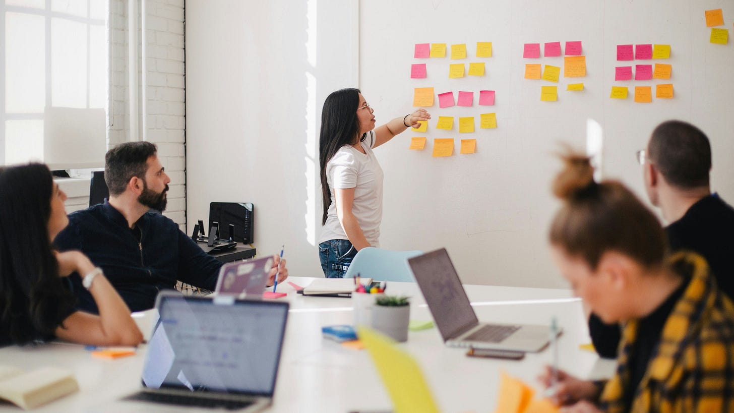 Team collaborating in a modern office space, with a woman presenting ideas on a whiteboard covered in colorful sticky notes while colleagues take notes and work on laptops.