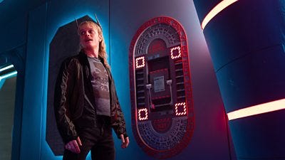 A man with horns stands beside a panel in a spaceship, looking shifty