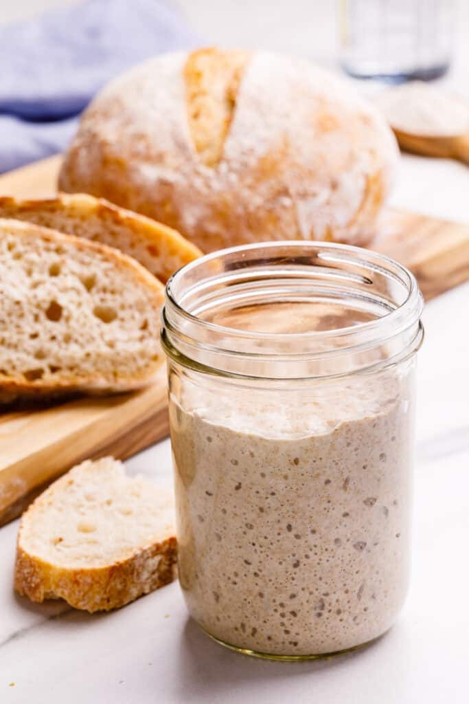 A glass jar of sourdough starter next to a loaf of sourdough bread.