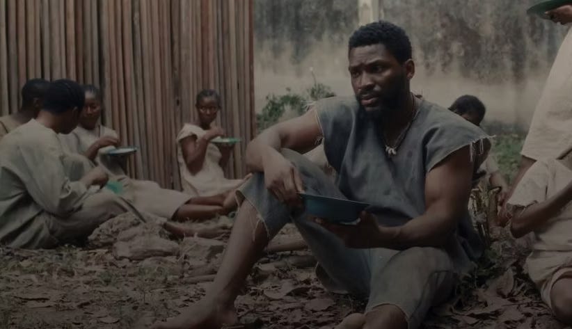 The image shows a man sitting on the ground, leaning forward with his elbows on his knees, holding a small bowl. He is wearing simple, worn clothing and a necklace with what appears to be teeth or claws. In the background, several other people are visible, also sitting on the ground with bowls, against a backdrop of wooden structures and greenery. The overall setting appears to be outdoors, with dirt and some foliage on the ground The image shows a man sitting on the ground, leaning forward with his elbows on his knees, holding a small bowl. He is wearing simple, worn clothing and a necklace with what appears to be teeth or claws. In the background, several other people are visible, also sitting on the ground with bowls, against a backdrop of wooden structures and greenery. The overall setting appears to be outdoors, with dirt and some foliage on the ground