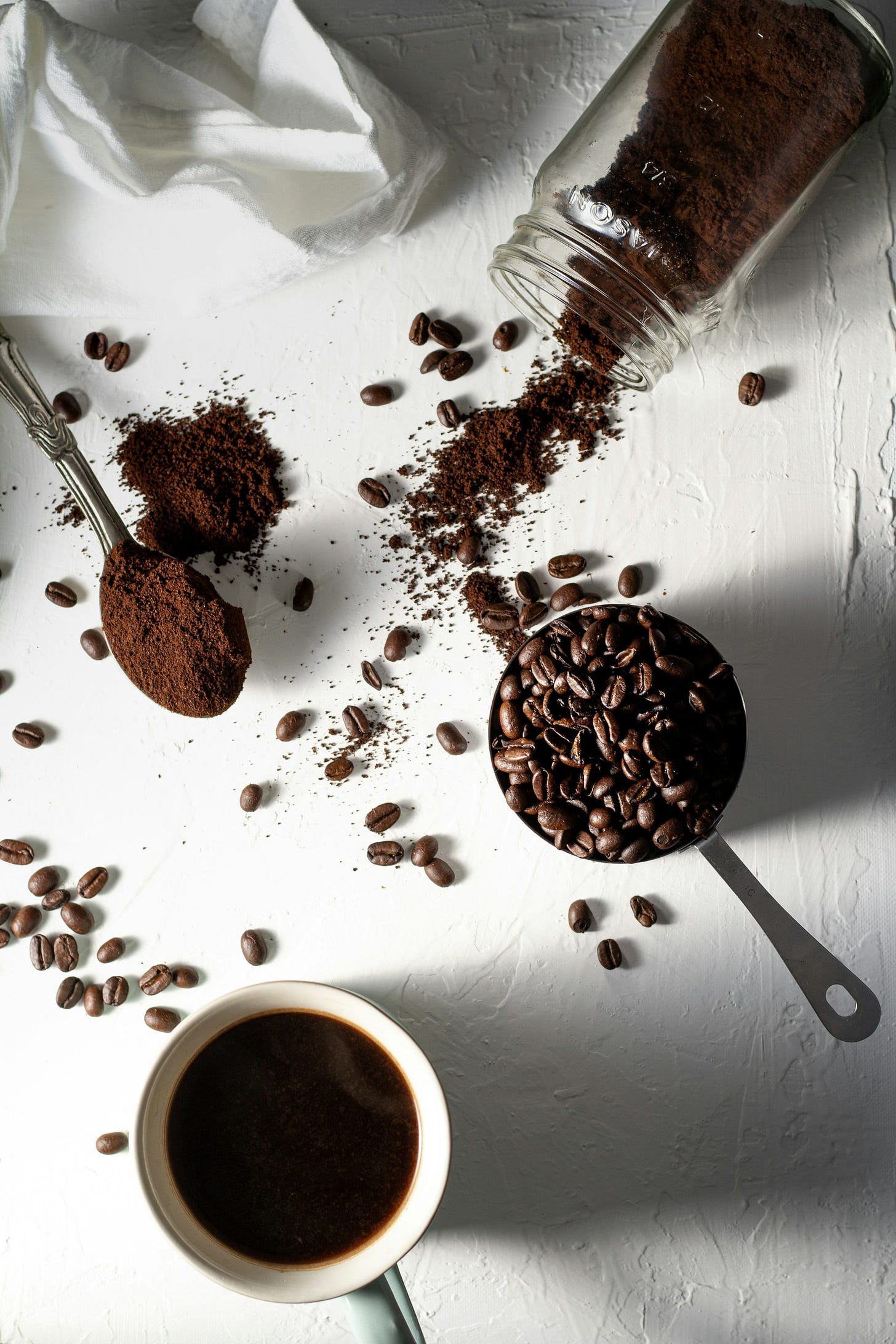 A white painted surface on which rest a tipped glass jar spilling ground coffee, a metal measuring cup holding coffee beans, a teaspoon with ground coffee heaped on it and spilled over, loose coffee beans, a white napkin skrunched at the top left, and a white cup of black coffee at the bottom.
