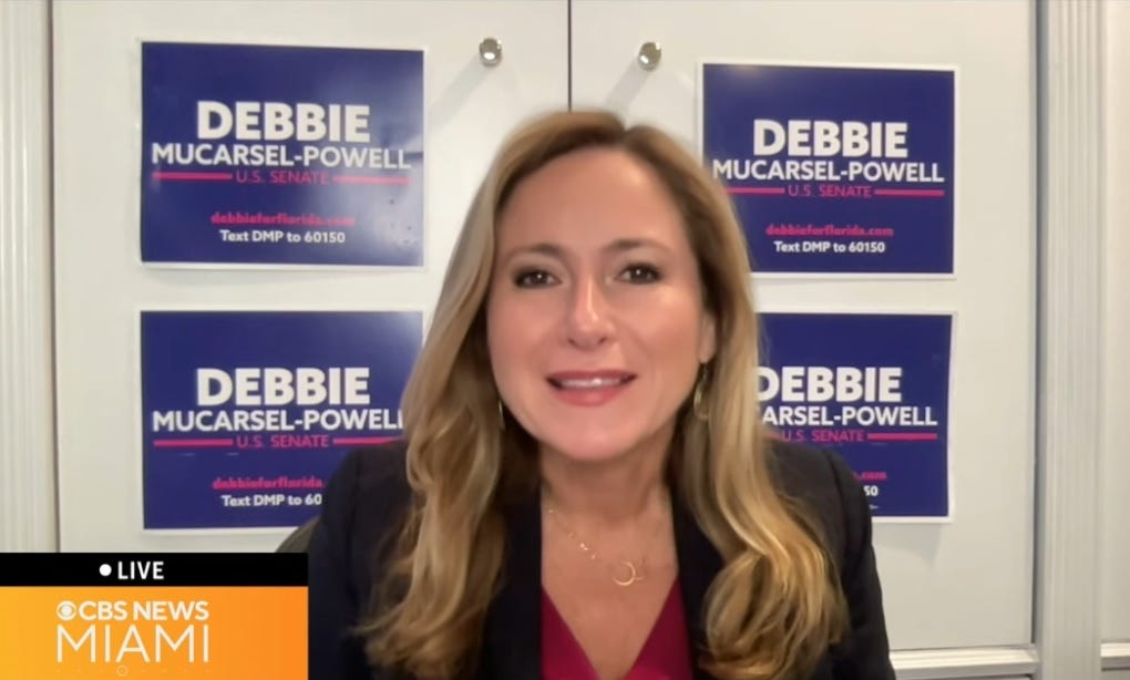 Debbie Mucarsel-Powell in a local TV news interview, standing in front of several of her own campaign signs on a wall Debbie Mucarsel-Powell in a local TV news interview, standing in front of several of her own campaign signs on a wall