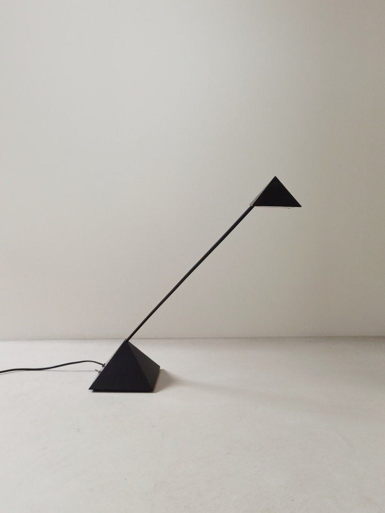 May include: A black triangular floor lamp with a long, slender arm extending from a triangular base to a smaller triangular shade. The lamp is minimalist and modern in design, suitable for contemporary interiors. Its sleek lines and simple geometry create a clean, uncluttered aesthetic. The matte black finish adds a touch of sophistication. The lamp is shown against a plain white background, highlighting its form and shape. This stylish lamp would be a great addition to any modern home or office.