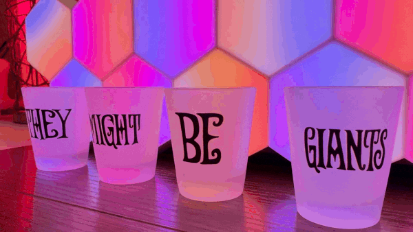 Four They Might Be Giants shot glasses arranged in a row spelling out THEY MIGHT BE GIANTS, backlit by shifting pink, purple, and orange honeycomb lights.