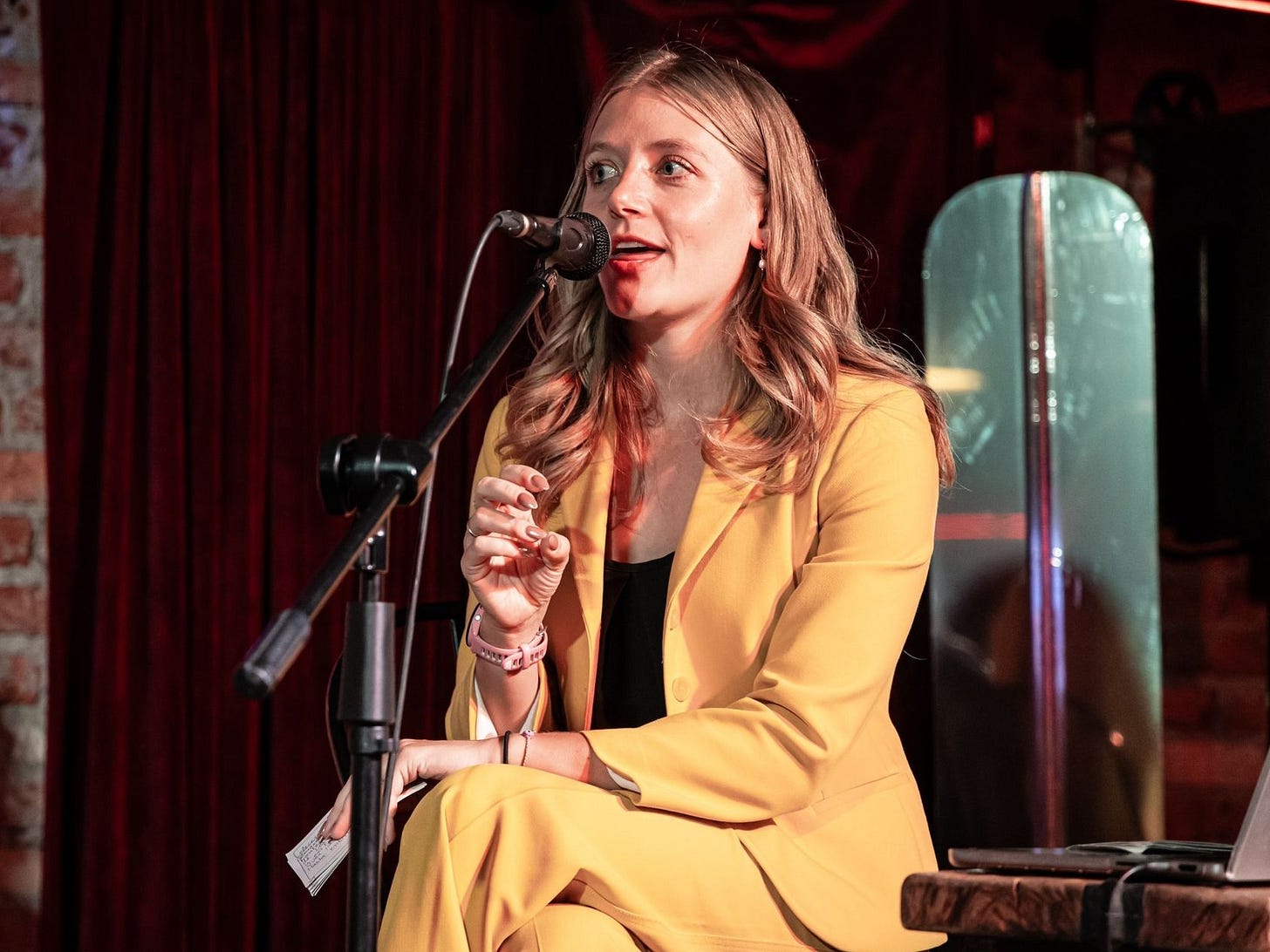 A young white woman sits on a stage with a microphone in front of her. Her mouth is open as if she is speaking into the microphone, and she is wearing a yellow pantsuit with a black shirt. She has shoulder-length blond hair.