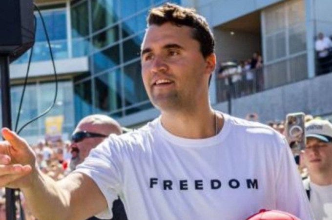 Charlie Kirk wearing a white T-shirt with the word "FREEDOM" in black capital letters. He is outdoors, raising his right hand, with a crowd and modern building visible in the background.
