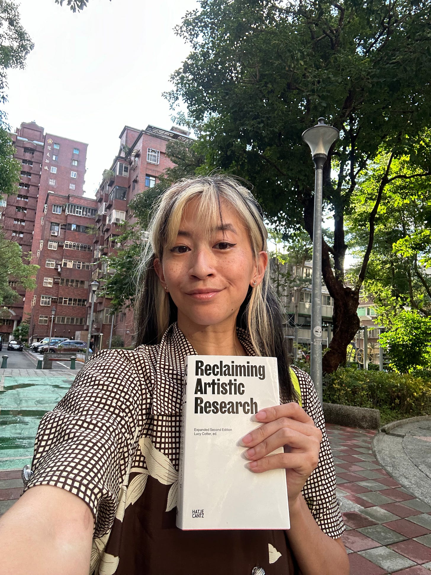 Yo-Yo in a city park holding up the book Reclaiming Artistic Research
