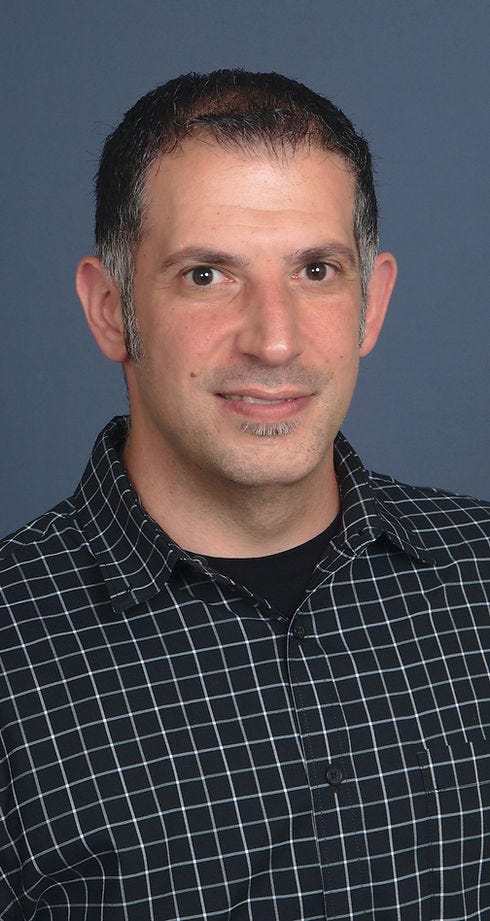 A man with short hair and a slight beard is smiling, wearing a black and white checkered shirt against a blue background.