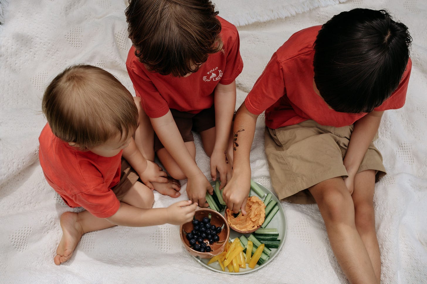 3 young boys in red t shirts sitting on a white blanket eating a veggie plate 3 young boys in red t shirts sitting on a white blanket eating a veggie plate