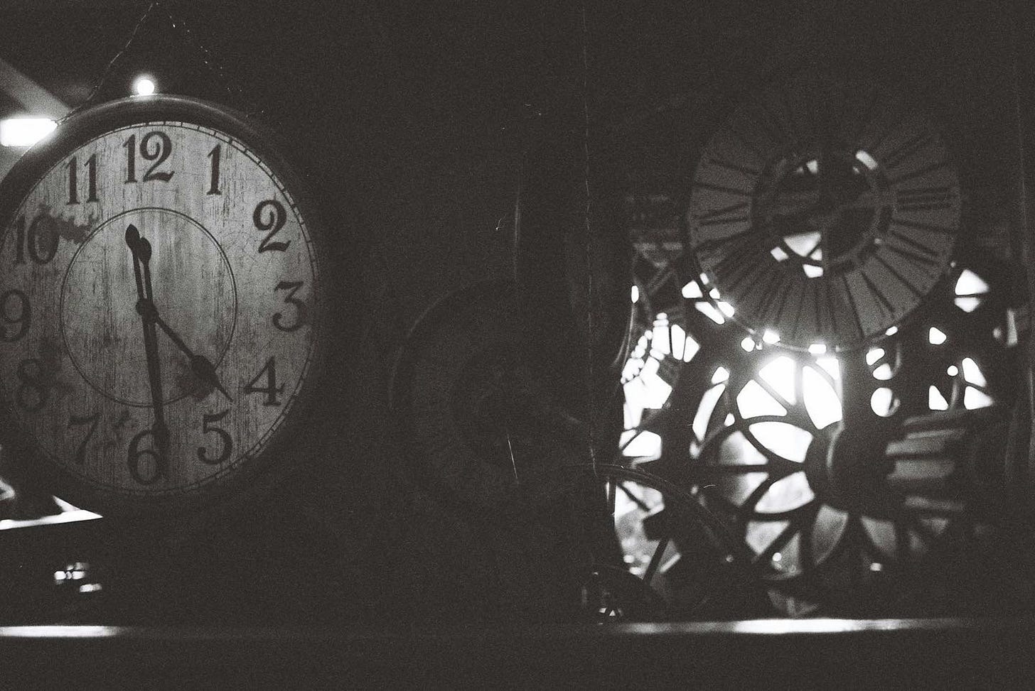 Black-and-white photo of antique clock faces and exposed gears; a large weathered wall clock reads about 6:55 while backlight shines through the machinery Black-and-white photo of antique clock faces and exposed gears; a large weathered wall clock reads about 6:55 while backlight shines through the machinery