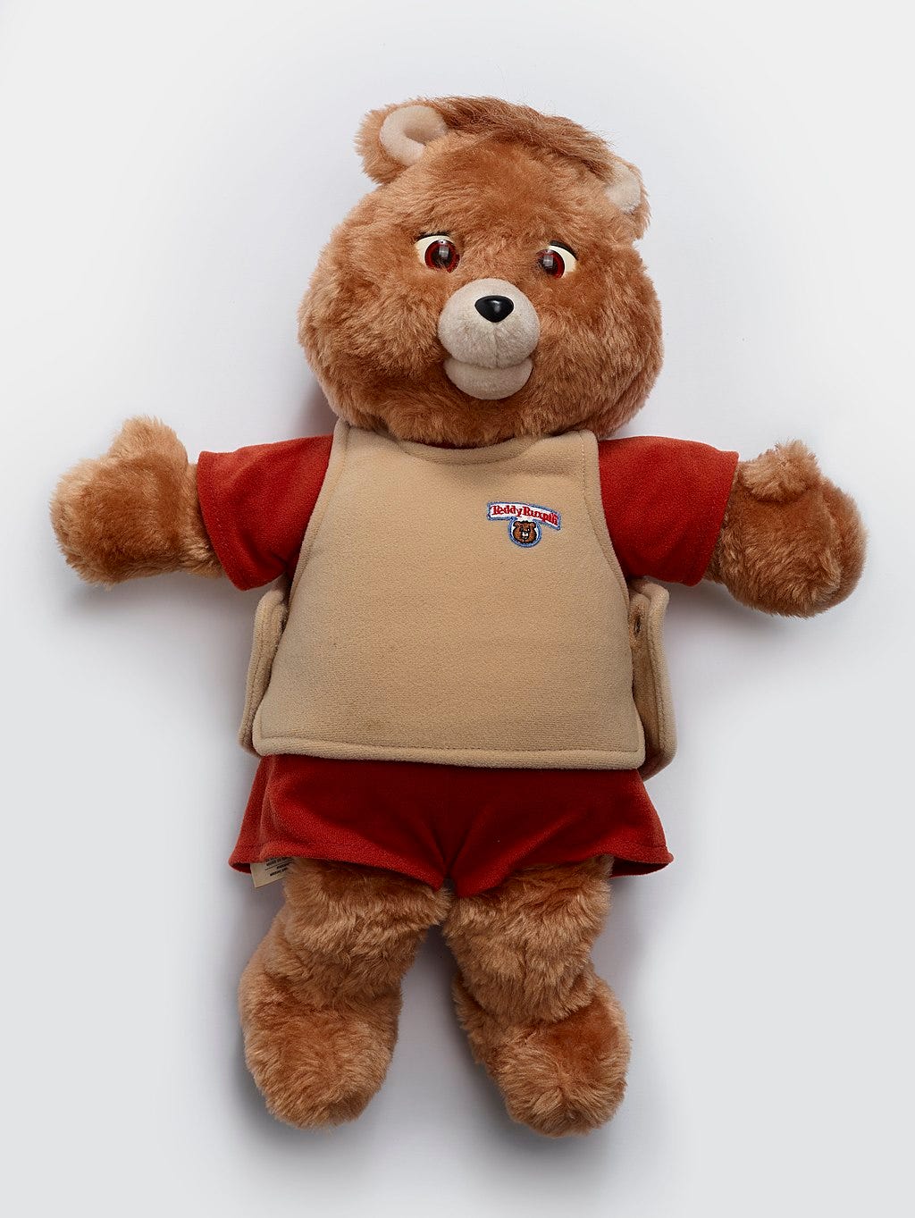 A high-resolution, full-body photograph of a 1985 Teddy Ruxpin animatronic bear. The toy features soft brown fur, a cream-colored muzzle, and large eyes with distinct amber irises. It is dressed in its signature outfit: a red short-sleeved shirt, a tan vest featuring the "Teddy Ruxpin" logo on the left chest, and matching red shorts. The bear is posed facing forward with its arms outstretched against a neutral, studio-lit background.