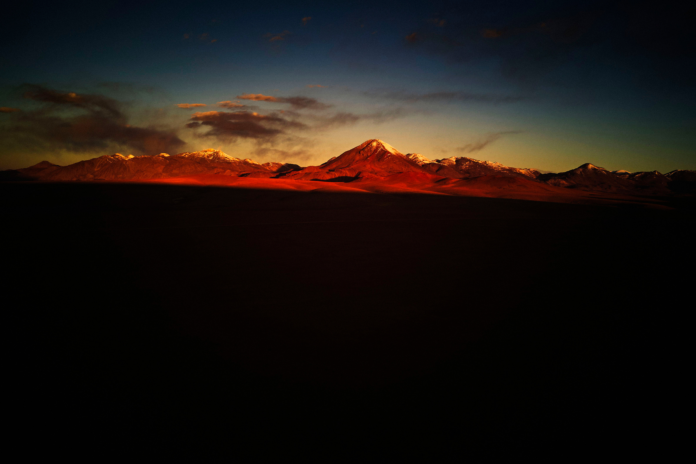 Sunrise or sunset light casts a warm, orange glow on distant snow-capped mountains under a dark sky with scattered clouds, while the foreground is mostly shadowed and dark.