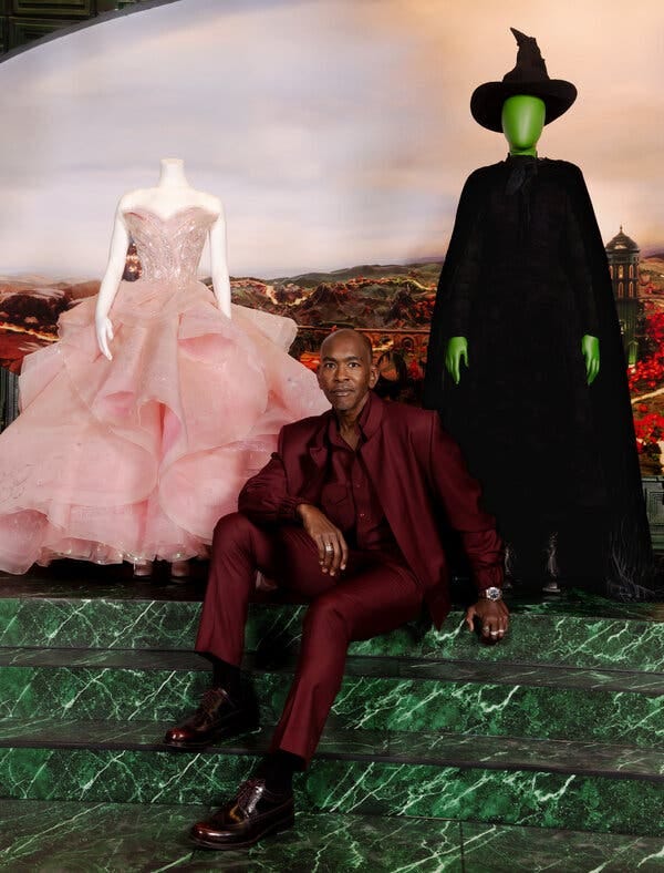 Paul Tazewell, who wears a shimmering burgundy suit and oxblood loafers, poses for a portrait on green steps. Arranged on mannequins behind him are two distinctive costumes from the “Wicked” movie: Glinda’s pink bubble dress and Elphaba’s black ensemble and pointy hat.