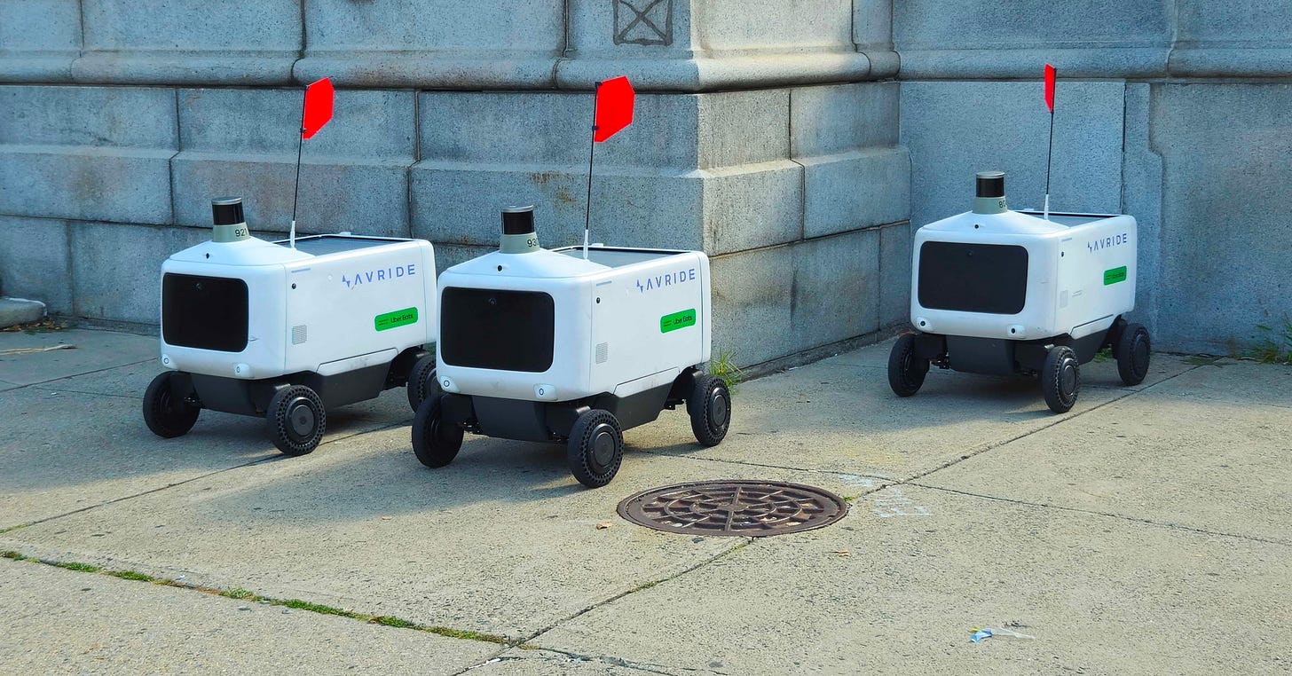 A photo of three Uber Eats autonomous delivery robots parked near a building during the day. A photo of three Uber Eats autonomous delivery robots parked near a building during the day.